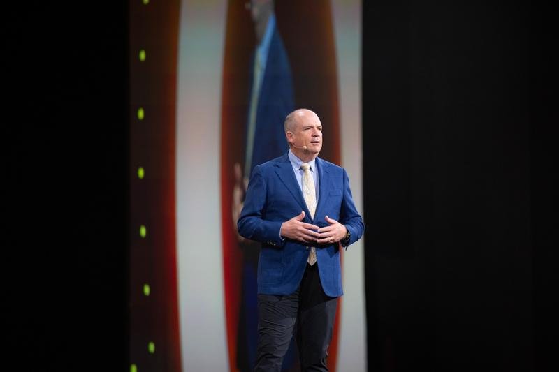 Основатель dōTERRA Грег Кук в синем пиджаке выступает на сцене Leadership Retreat