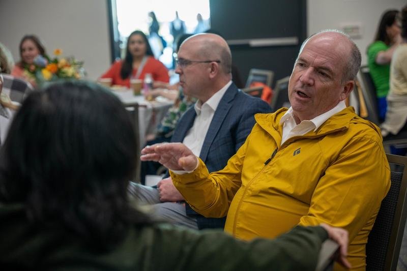 Грег Кук в жёлтой куртке увлечённо говорит с участниками за столом на Leadership Retreat в Сан-Диего